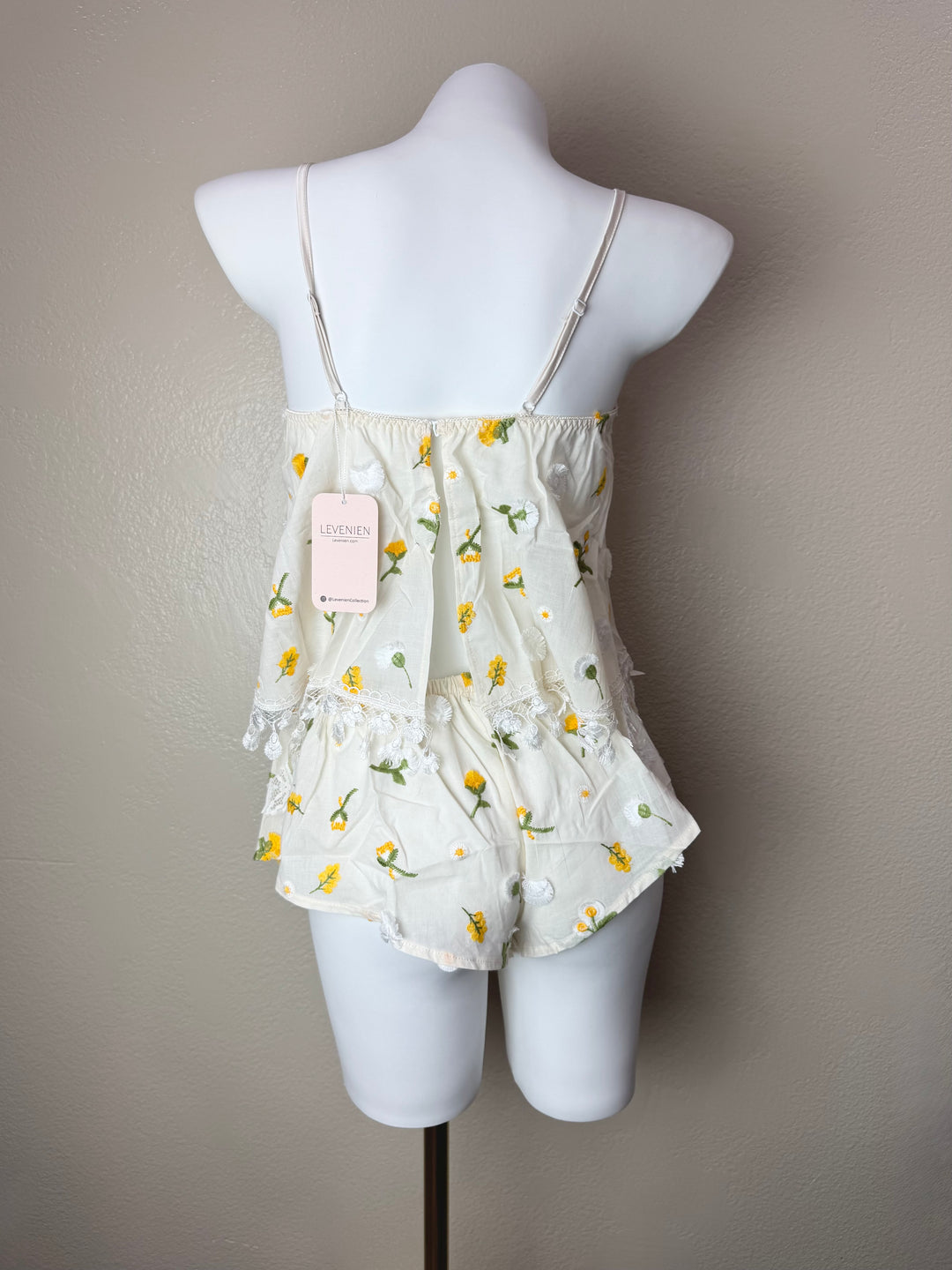 An off-white sleeveless camisole set with yellow and green floral embroidery, displayed on a white mannequin torso against a neutral gray wall. A pink tag is attached to the neckline.
